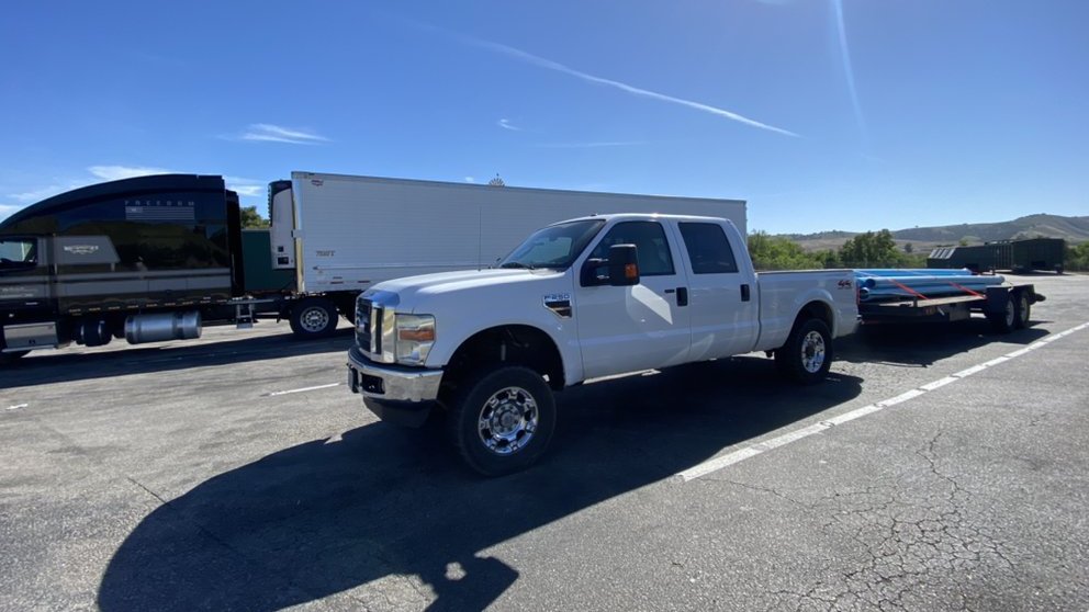 White Ford F-250 towing a flatbed trailer strapped with blue pipe, parked on an open lot under a clear sky