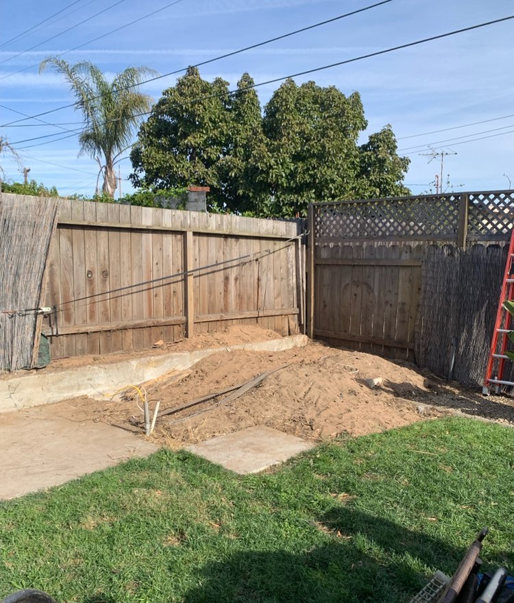 Backyard corner cleared to bare dirt along a wooden fence, lawn in front and a ladder by the gate